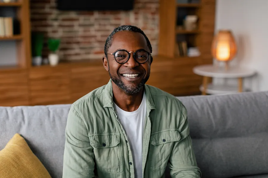 A middle aged black man in a green button up shirt sits on his couch smiling, happy with his treatment for andropause and low testosterone from Don D. Fisher, D.O. at The BEST Program in Fort Lauderdale.