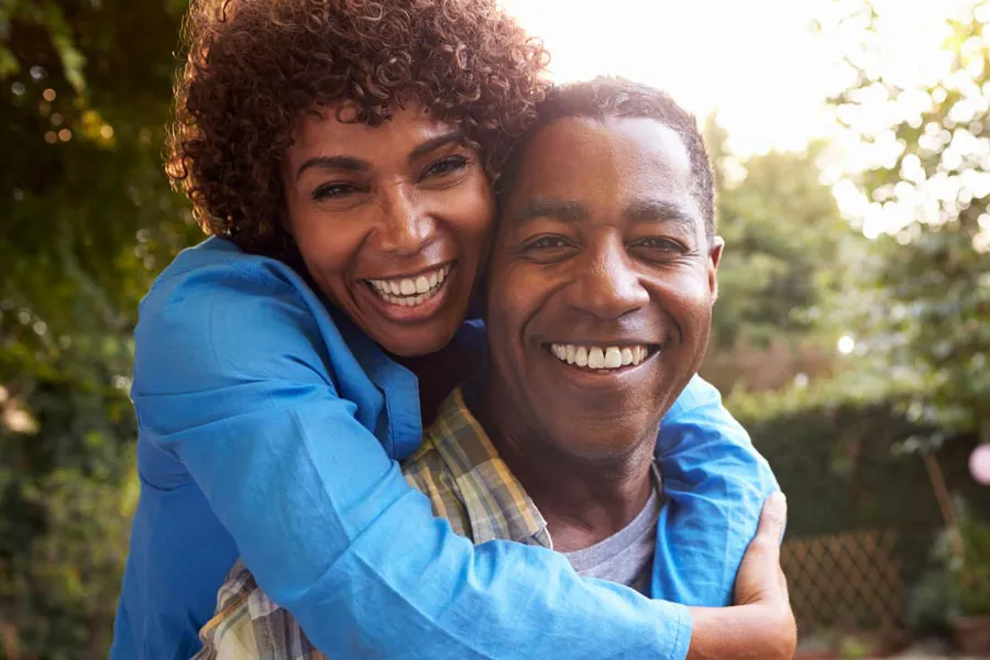 A middle-aged African American couple outside, the woman embracing the man from behind pressing her cheek to his, both are smiling due to successful Autoimmune Disorder treatment from Don D. Fisher, D.O. at The BEST Program in Fort Lauderdale.