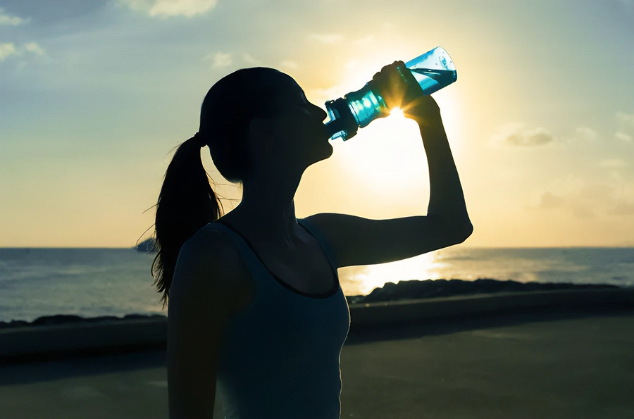 A woman in exercise clothes drinking from a water bottle on the beach, with the water and sunset in the background. Get chelation and detoxification treatment from [NAME] in [AREA].