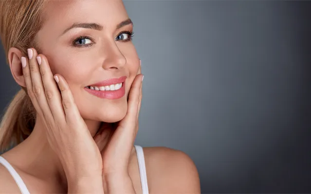 A middle-aged blonde woman with beautiful skin smiles and holds her face. Get facial treatments from Don D. Fisher, D.O. at The BEST Program in Fort Lauderdale.