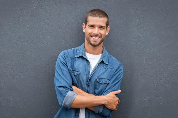 A man in a denim shirt stands smiling against a gray-blue wall, pleased with his testosterone hormone therapy from Don D. Fisher, D.O. at The BEST Program in Fort Lauderdale.