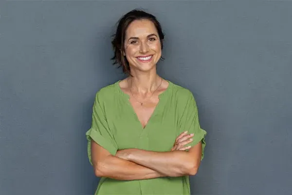 A woman in a green shirt stands smiling against a gray-blue wall, pleased with her hormone therapy from Don D. Fisher, D.O. at The BEST Program in Fort Lauderdale.