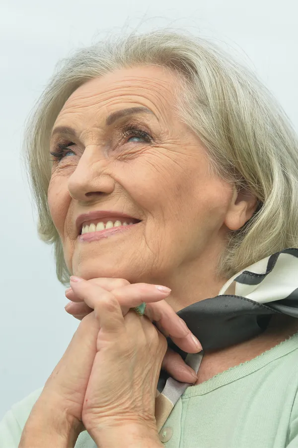 An older woman holds her hands together under her chin and looks up at a cool sky smiling. Get treatment for hot flashes and night sweats from Don D. Fisher, D.O. at The BEST Program in Fort Lauderdale.