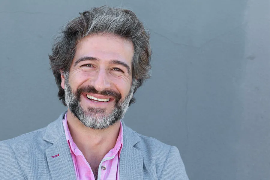 A smiling man with salt and pepper hair and beard wearing a pink shirt and gray blazer stands agains a gray wall. Get hypogonadism treatment from Don D. Fisher, D.O. at The BEST Program in Fort Lauderdale.