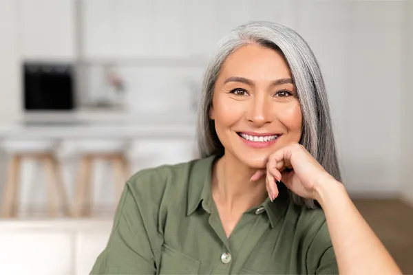 An asian woman with long gray hair and a green shirt sitting and smiling on her couch, happy with her menopause care from [NAME} in Fort Lauderdale.