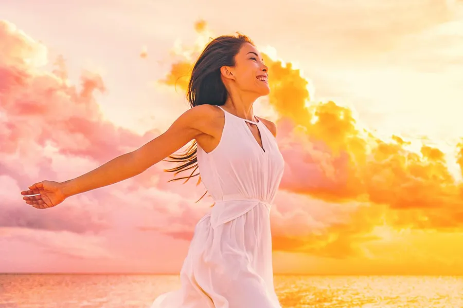 A woman in a thin white dress stretches out her arms joyously on the beach at sunrise. Get the Myers Cocktail IV treatment from Don D. Fisher, D.O. at The BEST Program in Fort Lauderdale.