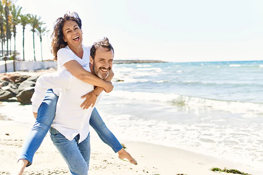 A woman piggybacks on her husband as the middle-aged couple have fun at the beach. Get NAD Therapy from [NAME] in [AREA].
