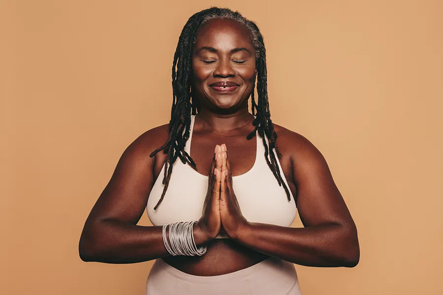 An older African American woman confidently posing against a warm background smiling with hands in a prayer pose. Don D. Fisher, D.O. at The BEST Program treats obesity using functional medicine in Fort Lauderdale.