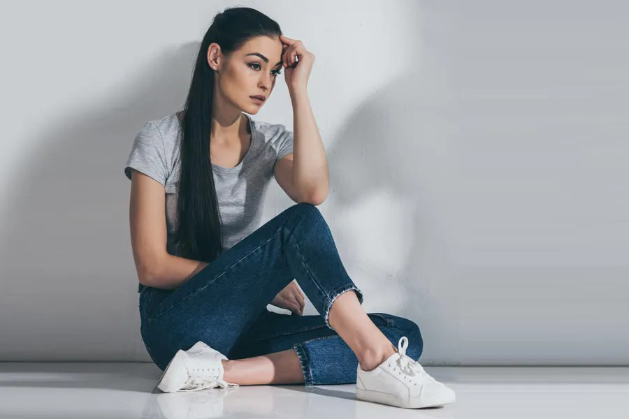 A young brunette woman in a gray t-shirt and jeans sits against a gray wall with her head resting on her hand and a distressed expression. Get help for Polycystic Ovarian Syndrome (PCOS) from Don D. Fisher, D.O. at The BEST Program in Fort Lauderdale.