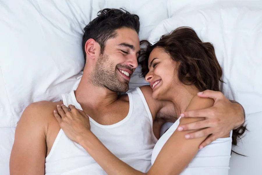 A laughing man and woman in undershirts lie in each other's arms on a white bed, representing successful PT141 sexual wellness therapy for men from Don D. Fisher, D.O. at The BEST Program in Fort Lauderdale.