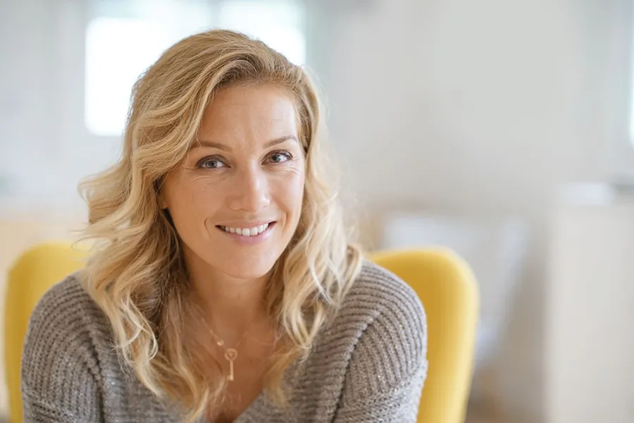 A blonde woman wearing a a gray sweater sits inside in a yellow chair, smiling and happy with her perimenopause treatment from Don D. Fisher, D.O. at The BEST Program in Fort Lauderdale.