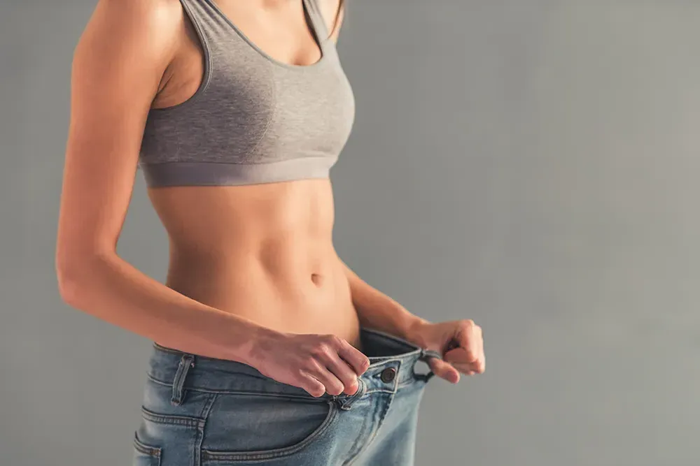 A woman pulls too-big jeans away from her waist to show weight loss. Schedule Tesamorelin peptide treatment from Don D. Fisher, D.O. at The BEST Program in Fort Lauderdale.