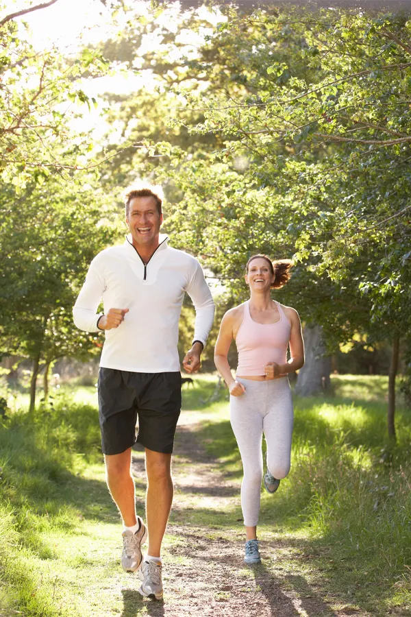 A middle-aged couple jog on a forest trail. Get Tesamorelin peptide treatment from Don D. Fisher, D.O. at The BEST Program in Fort Lauderdale.