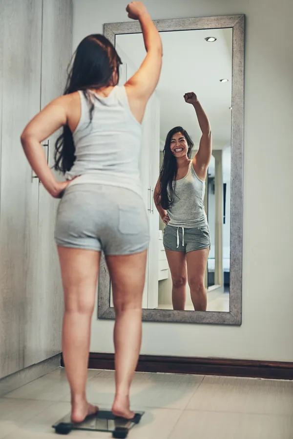 A woman in workout clothes stands on a scale looking in the mirror and pumping her hands in celebration of her successful Tirzepatide treatment from Don D. Fisher, D.O. at The BEST Program in Fort Lauderdale.